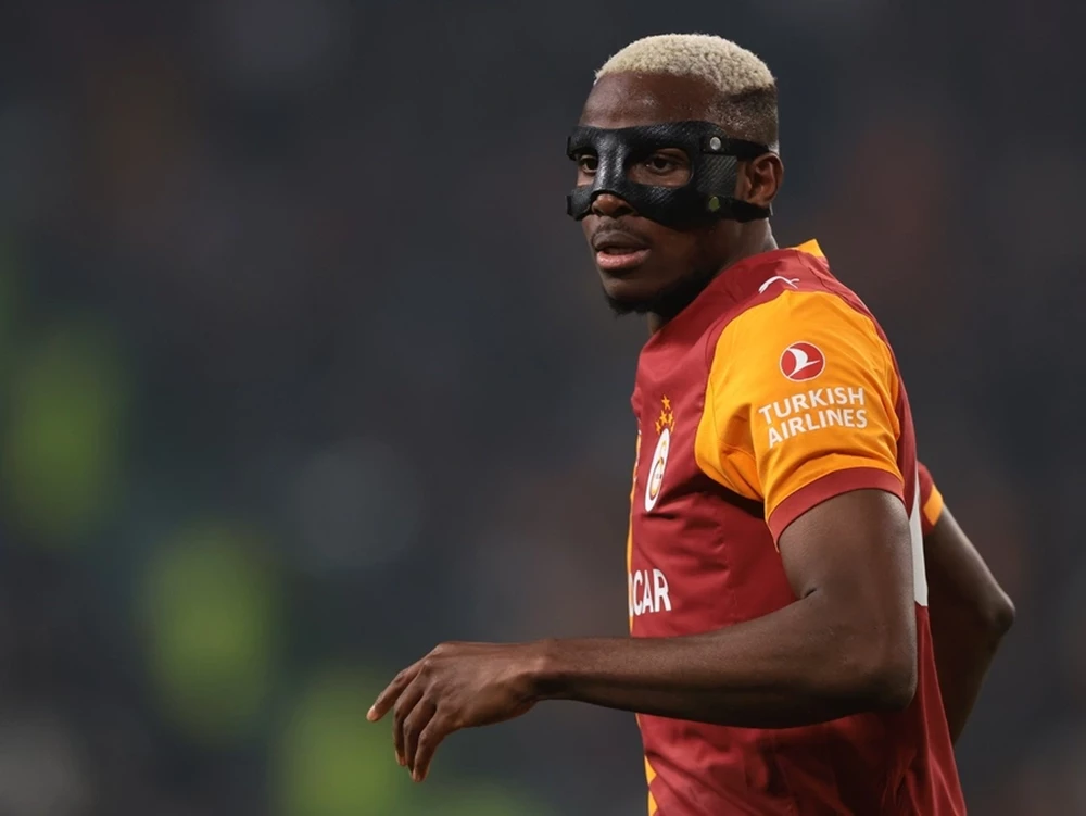Ein Spieler im rot-gelben Trikot mit einer schwarzen Gesichtsschutzmaske konzentriert auf dem Spielfeld. Erleben Sie Spitzenfußball live im Stadion.